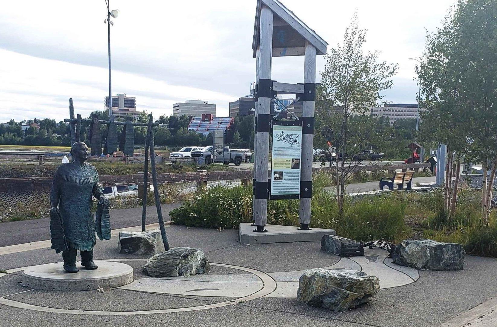 View of park with large stones on the ground, statue of a woman, trees, and Anchorage skyline in the background. 